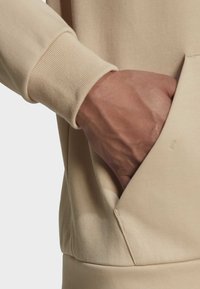 Mano colocada dentro del bolsillo frontal de un sudadera beige con puños acanalados, destacando la textura de la tela y los detalles de la costura.