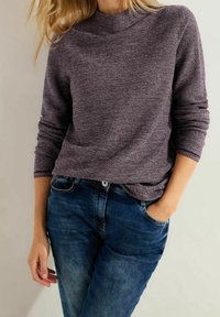 Lila strukturierte Langarmbluse mit hochgeschlossenem Kragen, kombiniert mit blauen Jeans. Die Bluse hat einen lässigen Schnitt und subtile Farbvariationen.