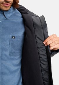 Zwarte jas met een gladde binnenkant en een fleece voering, gelaagd over een lichtblauwe corduroy shirt met een borstzak.