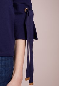 Tmavě modrý top s krátkým rukávem, ozdobený zlatými kroužky a dlouhými stuhami na rukávech. Hladká tkanina s jemným elastickým efektem.