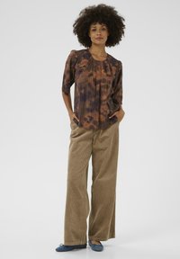Blouse tie-dye marron et orange avec des plis, associée à un pantalon en velours côtelé beige à jambes larges et des chaussures plates bleues, sur fond uni.