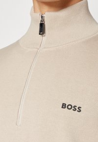 Pull beige zippé avec un col montant, texture côtelée, featuring un logo noir "BOSS" sur le côté gauche. Détail de fermeture éclair argentée.