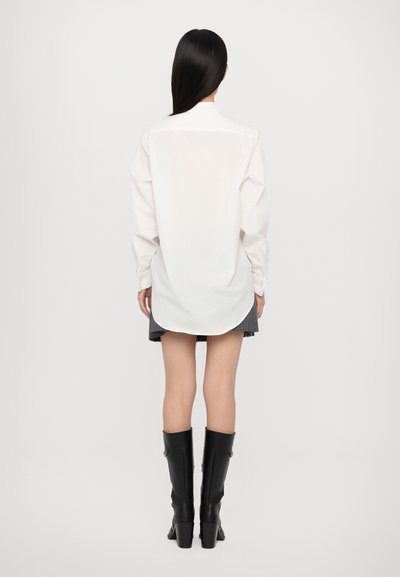 White oversized shirt with long sleeves, rounded hem, and button cuffs, paired with a gray pleated skirt and black knee-high heeled boots.