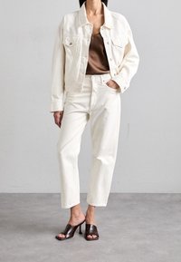 Bílá džínová bunda a kalhoty s hnědým topem. Outfit má volný střih, jednoduchý design a hladkou texturu. Styl doplňují hnědé boty na podpatku typu mule.