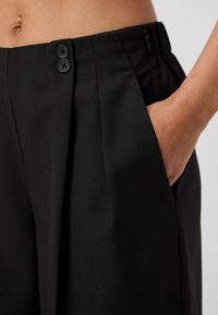 Black trousers with pleats, two black buttons at the waist, side pocket, and a smooth fabric texture. Zipper closure on the side.