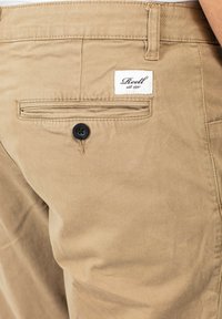 Khaki-Chinos aus weichem Baumwollgewebe, ausgestattet mit einer Gesäßtasche mit einem schwarzen Knopf und einem weißen Etikett mit schwarzem Text, das am Bund angenäht ist.