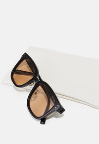Ochelari de soare supradimensionați, negri, cu lentile maro, având un design angular îndrăzneț, accente metalice și tampoane pentru nas transparente, expuși pe un pachet alb.