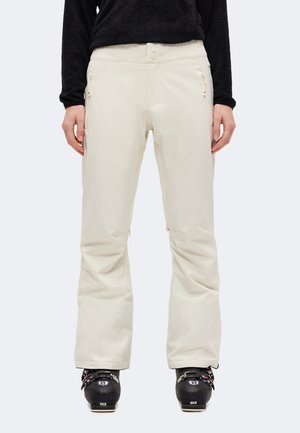 Un mannequin porte un pantalon de ski évasé couleur crème, un haut noir et des chaussures de ski noires sur un fond uni.