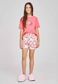 Różowy bawełniany t-shirt z grafiką jednorożca, zestawiony z białymi szortami ozdobionymi kolorowymi nadrukami jednorożców. Modelka ma na sobie różowe, puszyste kapcie w kształcie jednorożców.