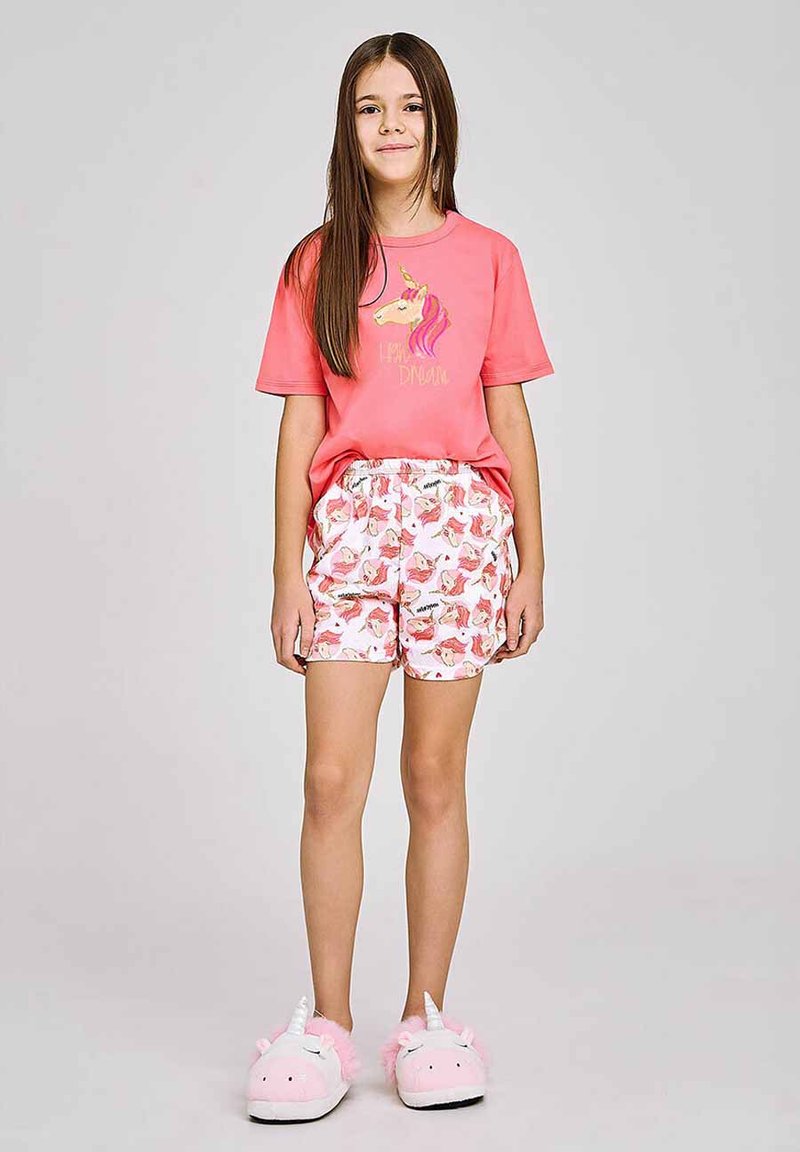 Różowy bawełniany t-shirt z grafiką jednorożca, zestawiony z białymi szortami ozdobionymi kolorowymi nadrukami jednorożców. Modelka ma na sobie różowe, puszyste kapcie w kształcie jednorożców.