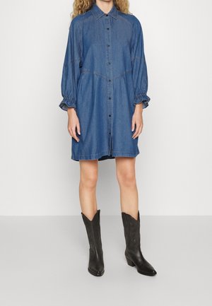 Kvinde iført en knælængde, blå denim kjole med knapper, lange puffede ærmer og sorte cowboystøvler, stående mod en ensfarvet baggrund.