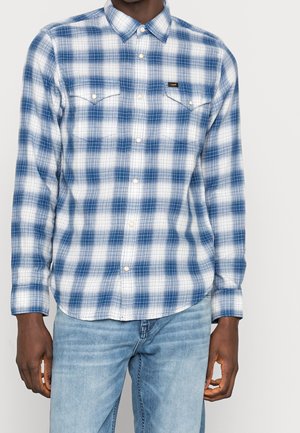 Mand iført en blå og hvid ternet skjorte med brystlommer og lyseblå jeans mod en ensfarvet baggrund.