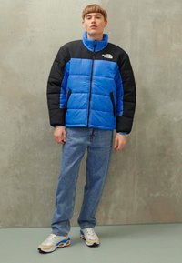 Polstret jakke i klar blå og sort, med lynlås foran, høj krave og to sidelommer. Båret med lyseblå jeans og sneakers.