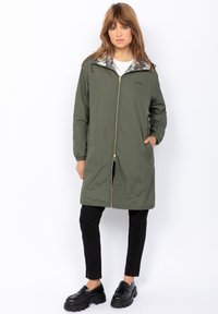 Donna che indossa una giacca lunga verde oliva con zip e polsini elasticizzati, pantaloni neri e mocassini neri spessi, in piedi su uno sfondo bianco.