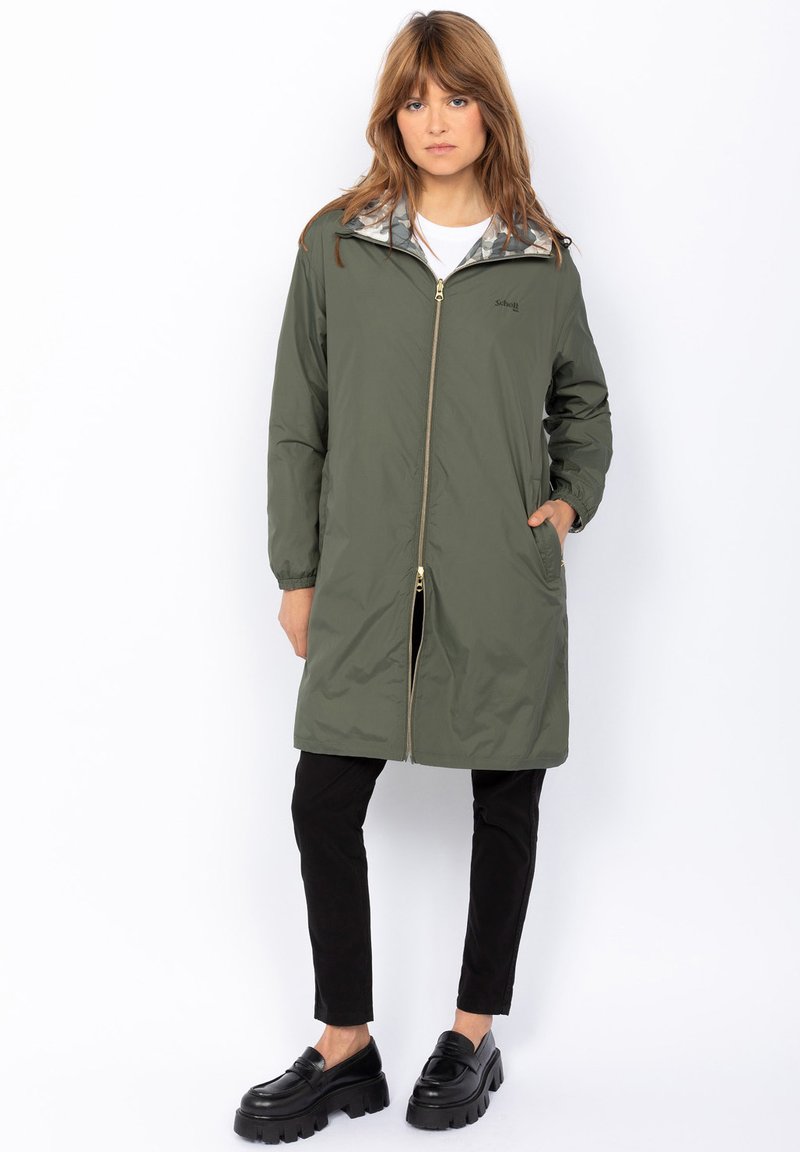 Donna che indossa una giacca lunga verde oliva con zip e polsini elasticizzati, pantaloni neri e mocassini neri spessi, in piedi su uno sfondo bianco.