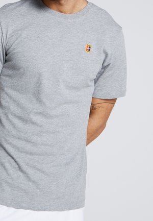 Homme portant un t-shirt gris clair à manches courtes avec un petit logo carré coloré sur le côté gauche de la poitrine et un tatouage partiellement visible sur l'avant-bras gauche.