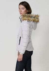 Világosszürke puffertáska műszőrmés kapucnival, barna és bézs árnyalatokban. Bordázott oldalsó panelek és fekete cipzáras zsebek jellemzik.