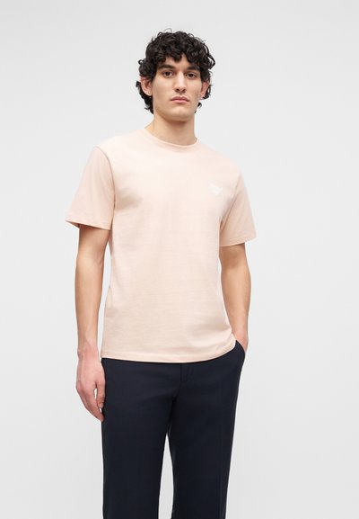 Νέος άνδρας με σγουρά σκούρα μαλλιά, που φοράει ανοιχτό ροζ κοντομάνικη μπλούζα και σκούρο παντελόνι, στέκεται με το ένα χέρι στην τσέπη.