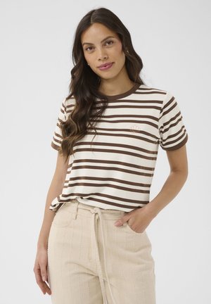 Pikkade laineliste juustega naine, kes kannab pruunvalgete triipudega t-särki ja beeži tekstuuriga pükse, millel on paelaga vöökoht, seisab ühe käega taskus.