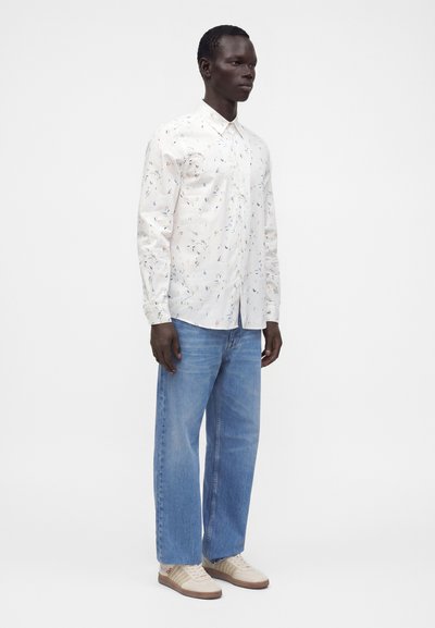 Un homme debout portant une chemise blanche à manches longues avec de petites taches colorées, un jean bleu et des baskets beiges sur un fond blanc uni.
