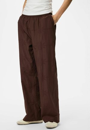 Osoba nosiaca voľné tmavohnedé nohavice s jemnými vyšívanými vzormi, vreckami, biely top a biele slip-on topánky.