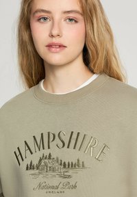 Oliwkowy sweter z ściągaczem, zdobiony srebrnym haftem z napisem "HAMPSHIRE" oraz sceną chaty z drzewami i napisem "Park Narodowy" poniżej.