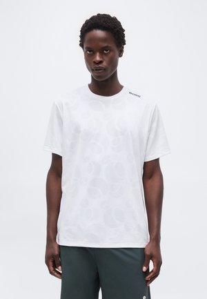 Ενήλικας με κοντά σγουρά μαλλιά, φοράει λευκό πουκάμισο με μοτίβα και κοντά μανίκια, καθώς και σκούρο πράσινο παντελόνι, στέκεται μπροστά σε απλό φόντο.