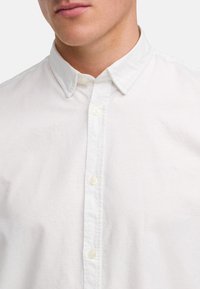 Camisa blanca de botones con cuello puntiagudo, que presenta pequeños botones y un discreto logo bordado en el pecho. Textura de tela de algodón suave.