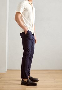 Korte mouwen crèmekleurig button-up shirt met een gestructureerd patroon, gecombineerd met navy slim-fit broek en zwarte instappers met een gouden accent.