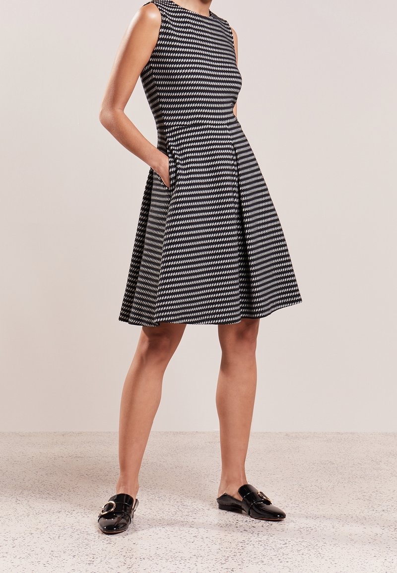 Femme portant une robe trapèze sans manches à motif pied-de-poule noir et blanc avec des poches, associée à des mocassins noirs brillants sans lacets.