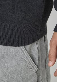 Gros plan sur une personne portant un pull en tricot noir et un pantalon en denim gris clair avec une poche latérale visible et une main reposant à côté.
