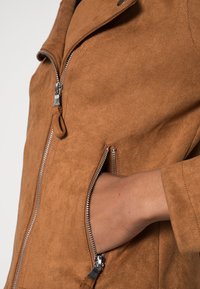 Veste en faux daim marron avec une fermeture éclair à l'avant et des poches latérales, présentant une texture douce et un design minimaliste. Quincaillerie de la fermeture éclair visible.