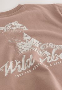 Camisa marrón claro con gráfico blanco de un leopardo reclinado y el texto "Wild Vibes" y "Look for adventure & have fun."