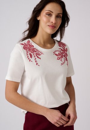 Femme portant une chemise blanche à manches courtes avec une broderie florale rouge sur les épaules et un pantalon rouge foncé, debout devant un fond uni.