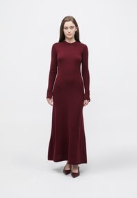 Bordowa dzianinowa sukienka z długimi rękawami, dopasowanym górnym partem i rozkloszowanym dołem. Gładka faktura i okrągły dekolt. Noszona z dopasowanymi szpilkami w spiczastym kształcie.