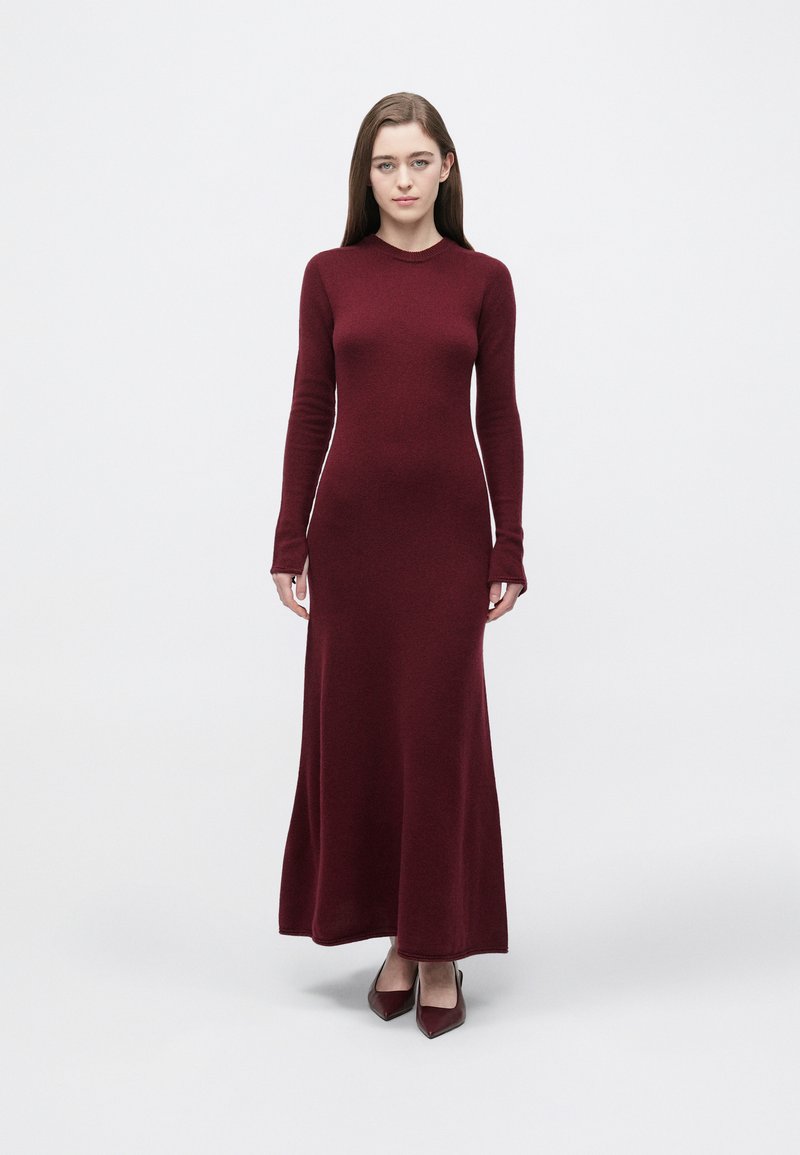 Bordowa dzianinowa sukienka z długimi rękawami, dopasowanym górnym partem i rozkloszowanym dołem. Gładka faktura i okrągły dekolt. Noszona z dopasowanymi szpilkami w spiczastym kształcie.