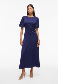 Žena v námořnickém midi šatech s nadýchanými rukávy a stříbrnými sandály na vysokém podpatku stojí na pozadí bílé zdi.