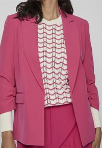 Rosa kavaj med en skräddarsydd passform och strukturerade axlar. Under den, en vit stickad topp med vågiga mönster i rosa accenter och ribbade manschetter.
