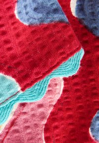 Tissu présentant un mélange de couleurs rouge, rose et bleu avec des motifs de tricot texturés. Comprend des bords festonnés en turquoise doux.
