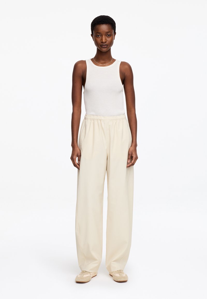 Femme noire debout, portant un haut blanc sans manches, un pantalon beige ample et des baskets beiges, sur un fond blanc uni.