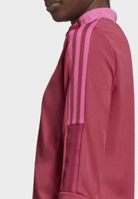 Camiseta deportiva rosa con cuello alto, tres rayas verticales rosas en los hombros y panel de malla texturizada en el lateral.