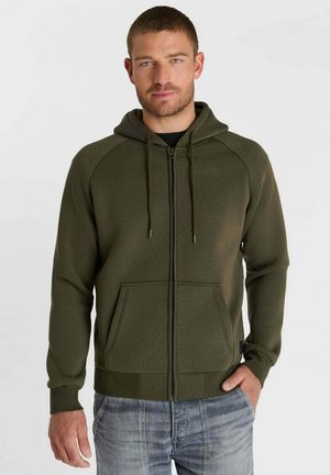 Man draagt een olijfgroene hoodie met rits en koordjes en een blauwe spijkerbroek, staand tegen een effen witte achtergrond.