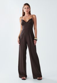 Žena s dlouhými rovnými vlasy, oblečená v tmavě hnědém bezramínkovém overal, s několika náramky na jedné zápěstí, stojící naboso.