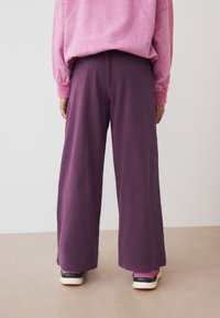 Kalhoty s širokými nohavicemi z měkké fialové látky s hladkou texturou. Nošeny s růžovou mikinou a kontrastními sportovními botami.