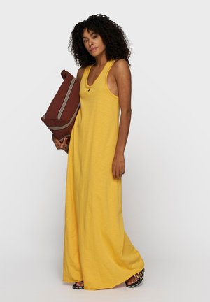 Kobieta z kręconymi włosami, ubrana w żółtą, bezrękawową sukienkę maxi, trzymająca dużą, brązową poduszkę w paski, stojąca na jednolitym tle.