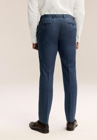 Pantalons bleus ajustés avec des poches arrière boutonnées et une texture lisse. Associés à des chaussures à lacets marron.