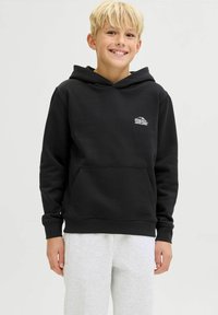 Blond chlapec sa usmieva, oblečený v čiernej mikine s malým bielym logom na hrudi a svetlošedých teplákoch, stojaci na jednolatom pozadí.