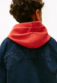 Granatowy sweter z teksturowanego materiału z czerwoną kapturem, z wyszywanym napisem "TOMMY" na plecach. Prążkowany materiał i kontrastujące detale kolorystyczne.