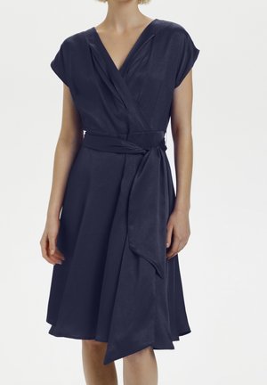 Woman wearing a navy blue wrap dress with short sleeves and a tied fabric belt at the waist, standing against a plain white background.