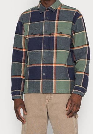 Uomo che indossa una camicia di flanella a quadri verde e blu navy abbottonata con tasche sul petto e pantaloni color beige su uno sfondo semplice.
